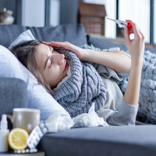 A woman with flu lies on the couch and looks at a fever thermometer.
