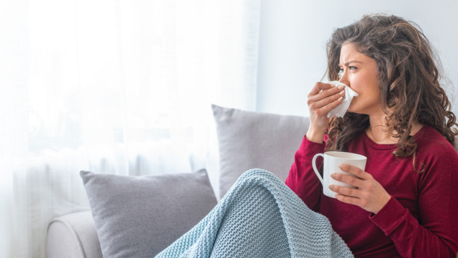 Woman with flu sits on the couch wrapped in a blanket and blows her nose.