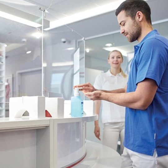 Man using hand disinfectant from a pump bottle at a counter in a medical or healthcare setting, with a woman in the background.