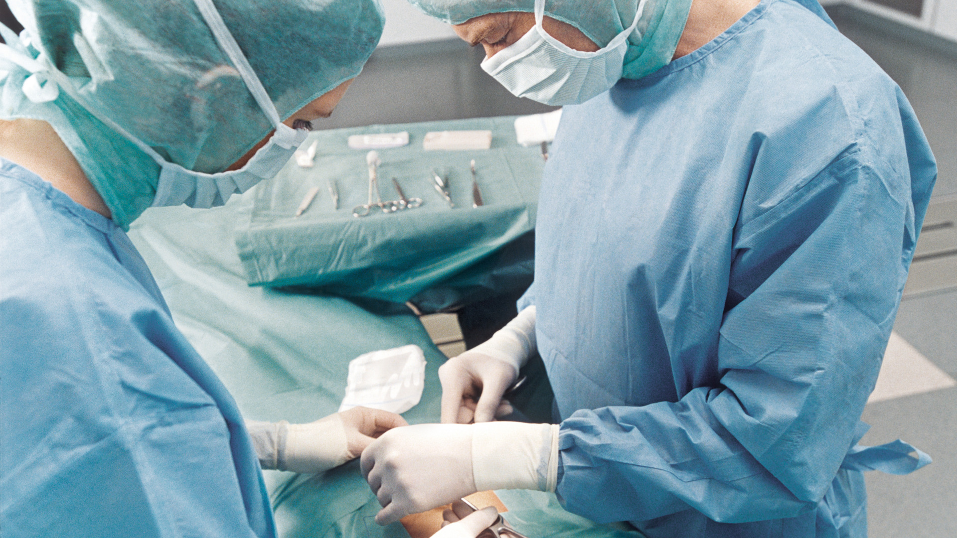  Two surgeons wearing blue surgical gowns and masks are focused on performing a precise procedure in a sterile operating room, with instruments on a tray.