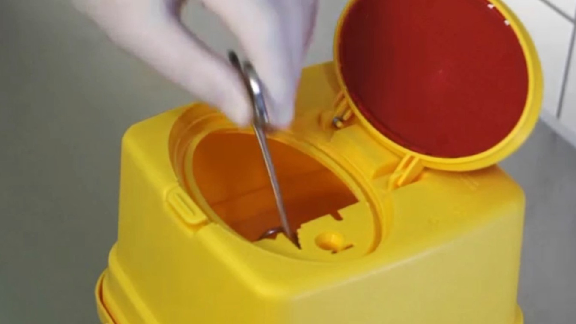 A gloved hand safely disposes of sharp medical instruments into a yellow sharps container with a red lid, highlighting efficiency and safety.