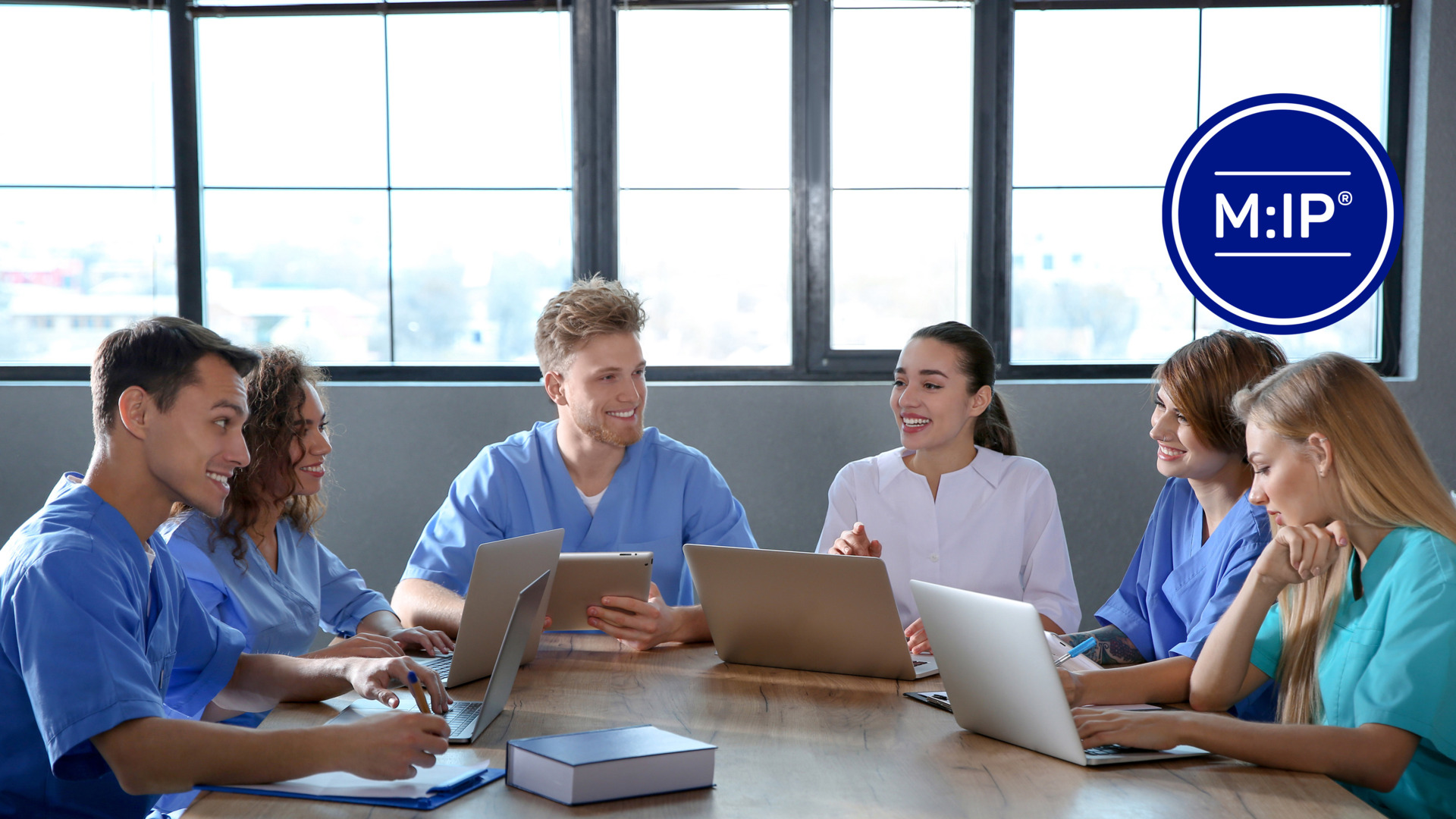 A group of healthcare professionals in uniforms gather around a table with laptops and tablets, engaged in an animated discussion in a modern office setting.