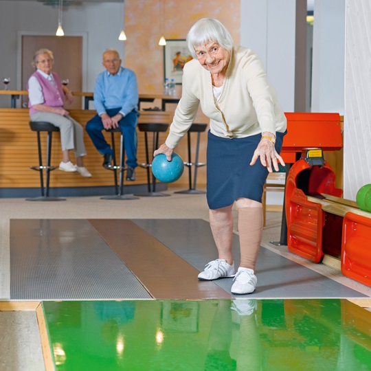 Seniorin wirft eine Kegelkugel. Im Hintergrund sitzen andere Senioren am Tisch