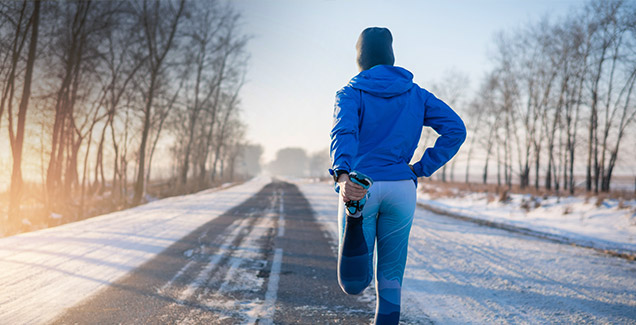 Rücken einer Frau mit winterlicher Sportbekleidung in einer Winterlandschaft. Sie zieht ihr Bein zum Rücken.
