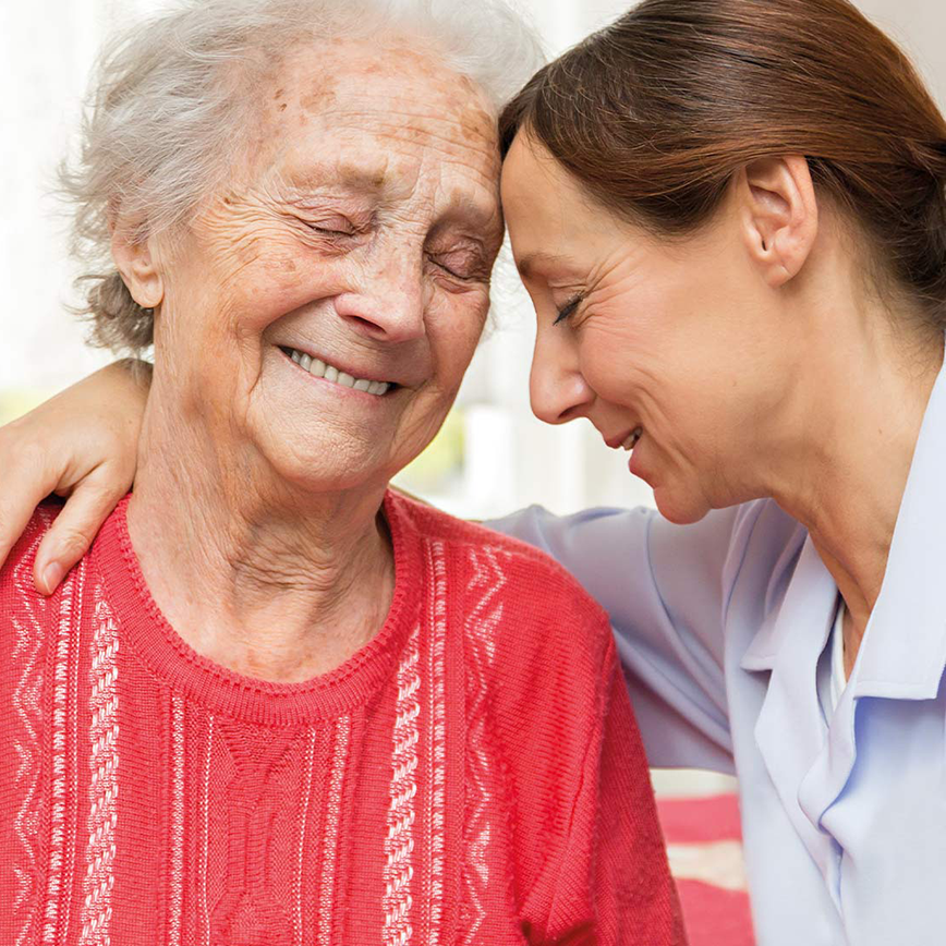 Pflegerin und Seniorin stecken lachend die Köpfe aneinander