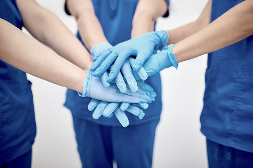 Personál nemocnice se zdravotnickými rukavicemi na rukou stojí v půlkruhu a mají položené ruce na sobě 