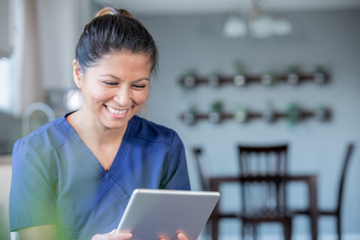 Lächelnde Mitarbeiterin des Gesundheitswesens in blauem Kasack, die in einem hellen, modernen Raum mit dekorierten Wänden sitzt und ein digitales Tablet benutzt.