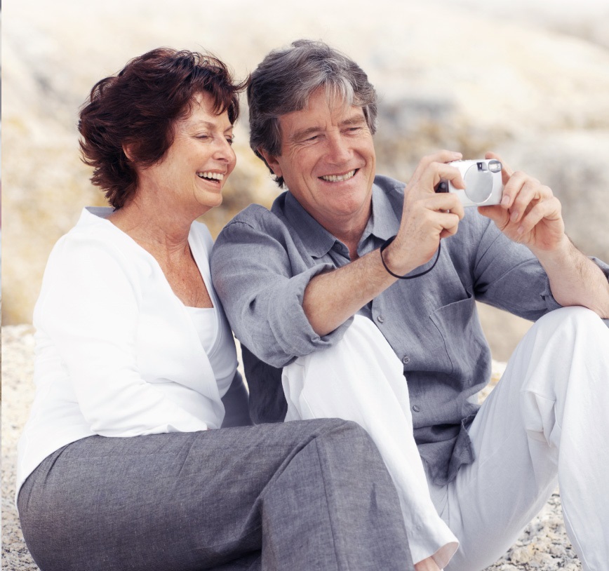 An elderly couple is sitting on rocky ground, while both of them are smiling into a camera, which the husband holds in front of them.