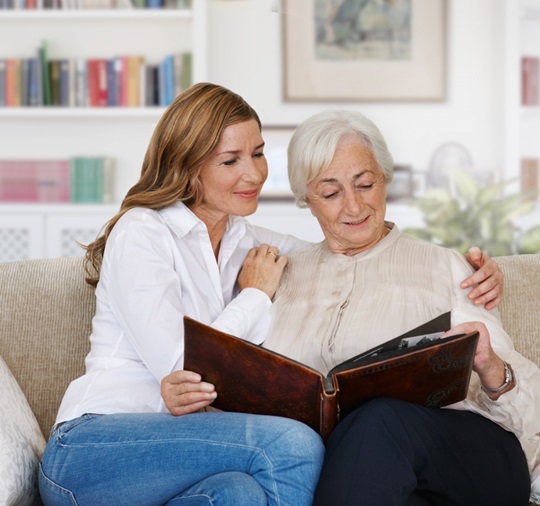 Une femme adulte est assise avec sa mère sur un divan dans un salon. Les deux femmes regardent joyeusement un album photo, tandis que la fille passe un bras autour des épaules de sa mère.