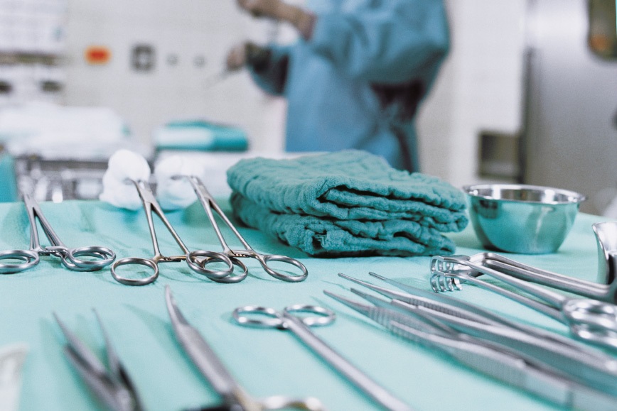 A close-up of a operation table with surgical instruments in an operation room; a doctor is standing behind the table.