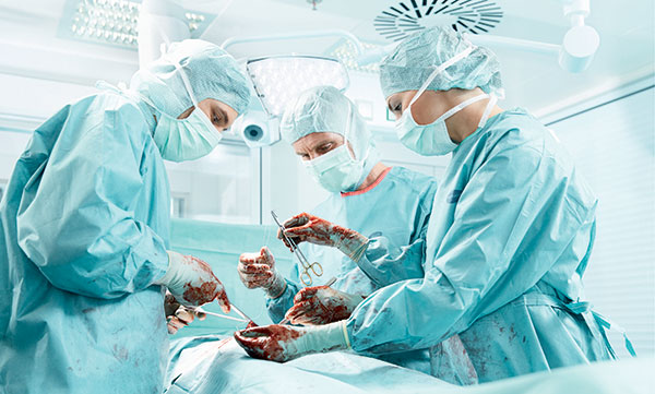 Detailní záběr na operační tým v průběhu operace ukazuje vybavení zdravotnického personálu současnosti, kukly, čepice, roušky, pláště, zástěry a samozřejmě i chirurgické rukavice