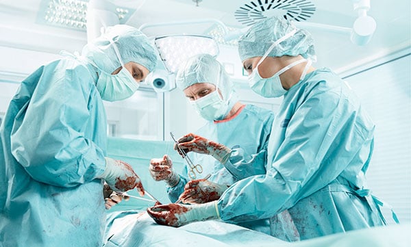Detailný záber na operačný tím v priebehu operácie ukazuje vybavenie zdravotníckeho personálu súčasnosti, kukly, čiapky, rúšky, plášte, zástery a samozrejme chirurgické rukavice