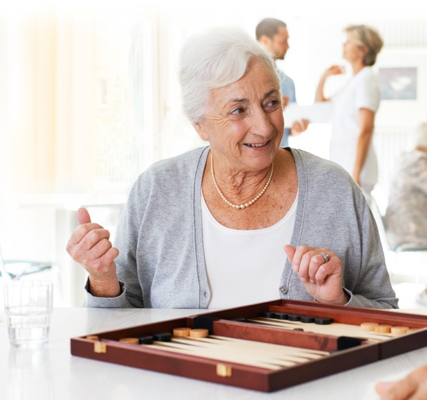 A happy-looking old lady is sitting at a table in a hospital room; in front of her is a board game. Two medical assistants standing far behind her and talking to each other. 