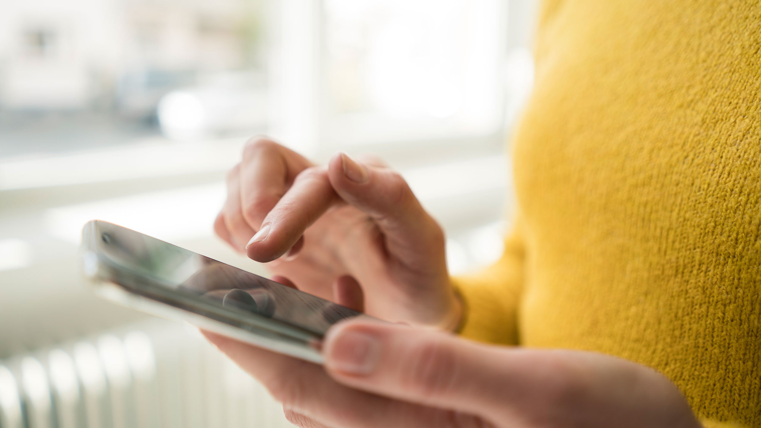 Close-up of hands holding and interacting with a smartphone, showcasing a yellow sweater in a softly lit room.