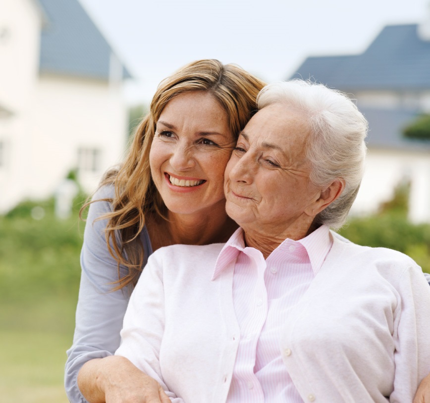 An adult woman is standing outside with her mother; both are smiling happily